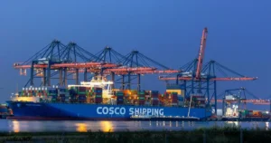 Dark blue night sky, large sea freight vessel holding colourful shipping containers alongside the quay, overhead gantry cranes loading and unloading cargo