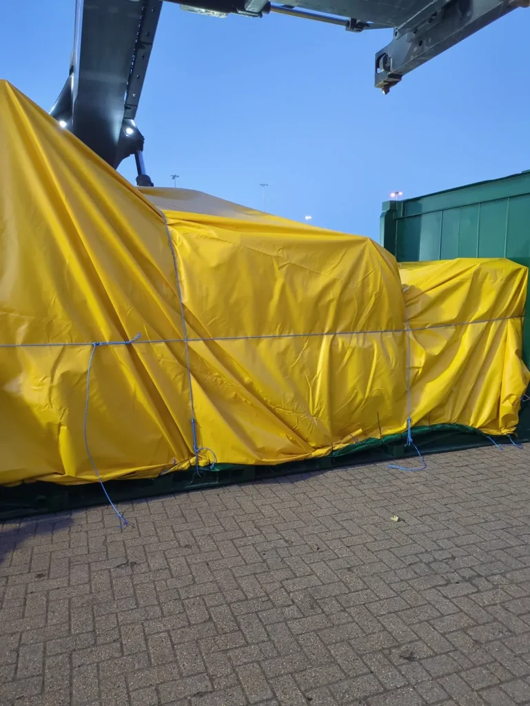 Side view of machinery covered with yellow sheeting, loaded on a 20' flat rack for out of gauge shipping to USA