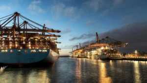 Dusk at a port, several sea freight vessels are docked alongside quay for loading and unloading, lights on the quay side and gantry cranes reflect off the water