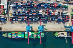 Ariel view of two sea freight vessels docked for unloading and loading by gantry cranes. Stacks of shipping containers mostly in blue alongside.