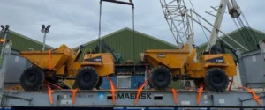 Two yellow dumper trucks, loaded on a grey flat rack container, lashed with bright orange strapping